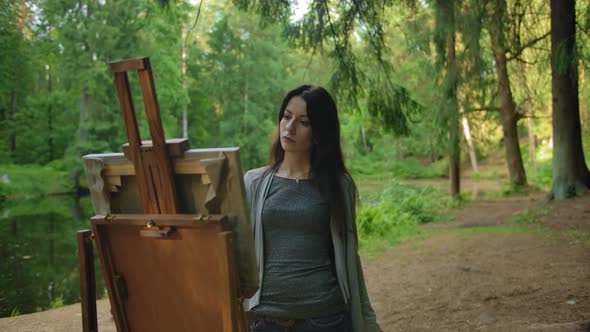 Beautiful Woman Artist in a Blouse and Jeans Paints a Landscape in a Park Near a Pond