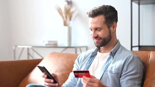 Cheerful Young Man Holds Credit Card and a Smartphone in Hands Shopping Online