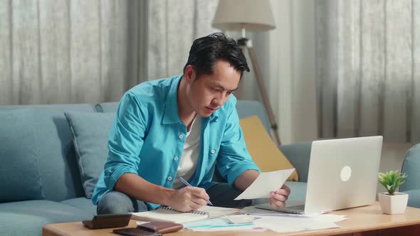 Asian Man With A Laptop Holding And Looking At The Bill While Recording The Expenses In Notebook