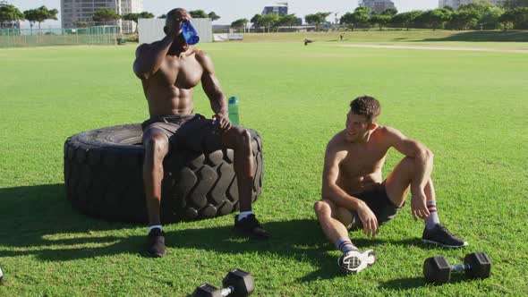 Two fit, shirtless diverse men resting, drinking water and bumping fists after exercising outdoors