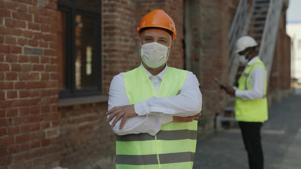 Builder in Mask Posing with Crossed Arms at Building Site