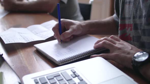 Close-up of man writing in notebook with laptop computer nearby