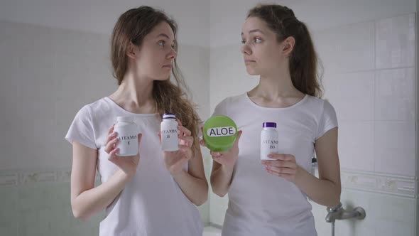 Two Beautiful Positive Twin Sisters Standing in Bathroom Holding Vitamin Bottles Smiling Looking at