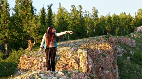 A Young Happy Woman with a Child in a Backpack on the Top of a Mountain Points with Her Finger