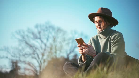 A Woman in a Stylish Hat Uses Her Phone While Sitting on the Grass in a Park