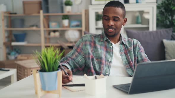 African American Guy Is Working at Home Using Laptop Typing Then Making Notes on Paper and Drinking