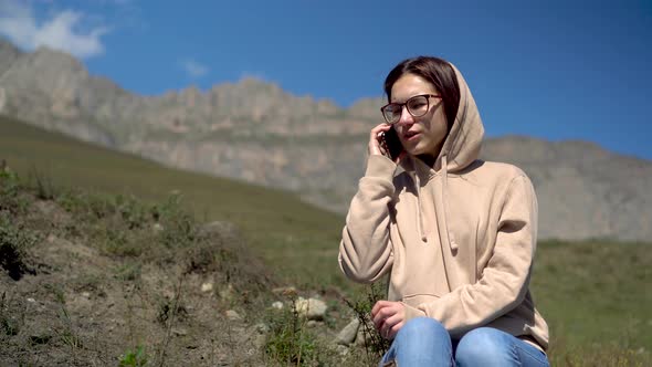 A Young Woman in a Sweatshirt Sits in the Mountains and Speaks on the Phone. The Girl Travels in the