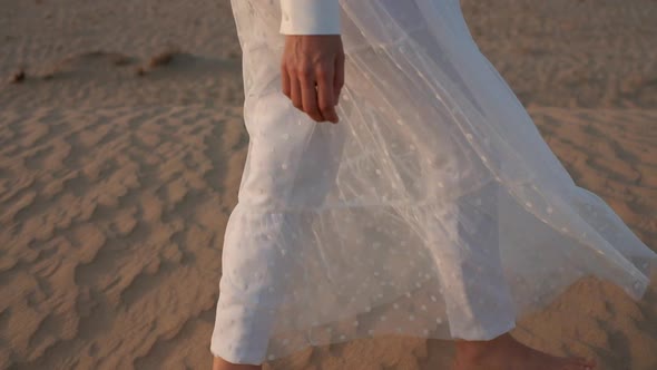 A Woman in a White Dress Barefoot is Walking Along the Desert Sand