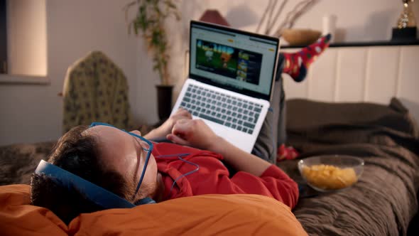 Young Man Lying on the Bed Watching Videos From the Laptop and Eating Chips