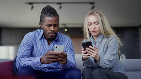 Medium Shot Portrait of Absorbed Interracial Couple Using Smartphones Sitting in Living Room