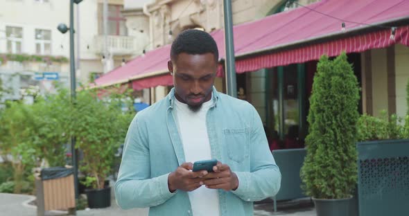 Black-Skinned Guy in Casual Clothes which Walking Near Street Cafe and Using His Mobile