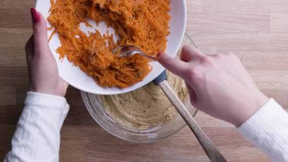 Hands Adding Grated Carrots with Tablespoon into Carrot Cake Dough. - Top Down Shot