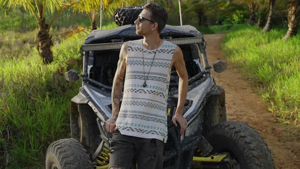 Male Wearing Sunglasses Leaning Against Bonnet Of Dirt Buggy And Folding Arms. Pedestal Down