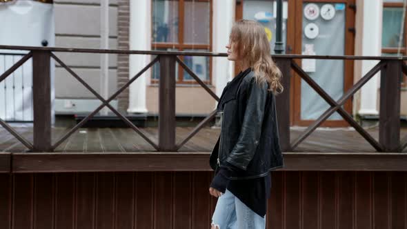 a Blonde in a Black Jacket Walks Against the Background of a Blurry House with a Veranda