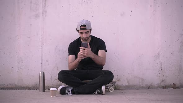 Man sitting on skateboard using phone and drinking coffee