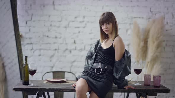 Portrait of Slim Gorgeous Young Woman in Black Dress and Leather Jacket Sitting on Wooden Table