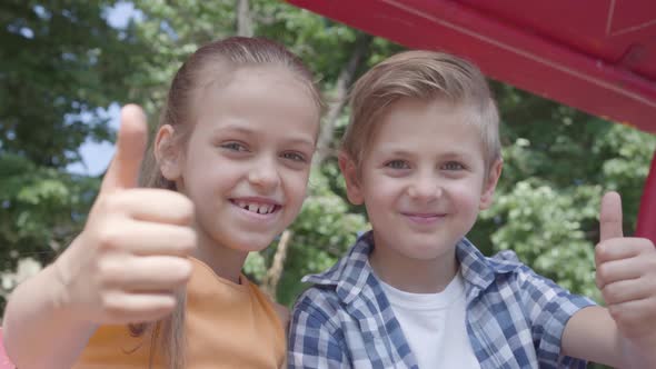 Cute Boy and Girl Showing Thumbs Up Sitting on the Swing Close Up in the Park, Looking Into Camera