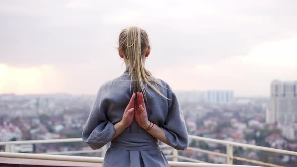 Blonde Woman Enjoys City View and Folds Her Hands Behind Back in a Namaste Gesture