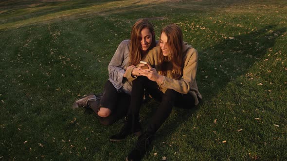 Twin teenage girls sitting on grassy hill looking at smartphone