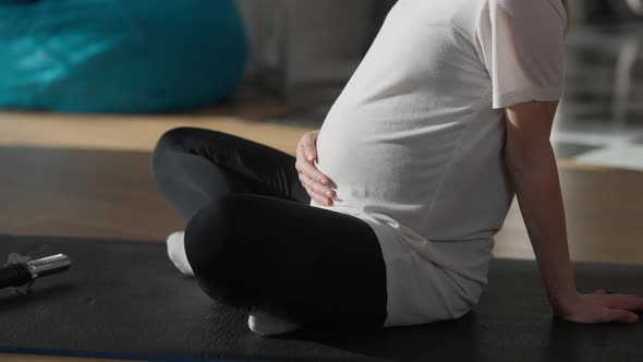 Unrecognizable Pregnant Sportswoman Caressing Belly Sitting on Exercise Mat Indoors