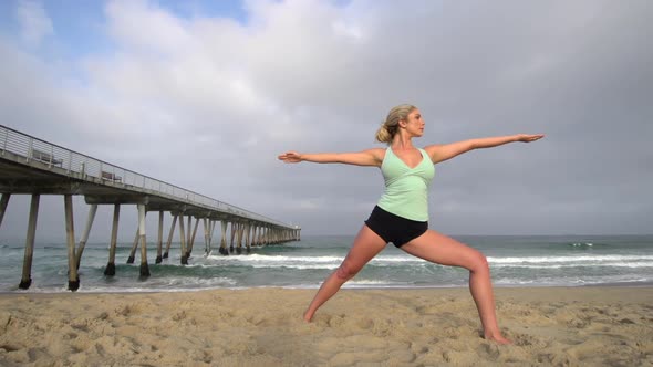 A young attractive woman doing yoga on the beach next to a pier.