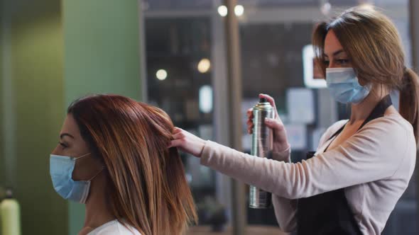 Female hairdresser wearing face mask spraying hairspray on hair of female customer at hair salon