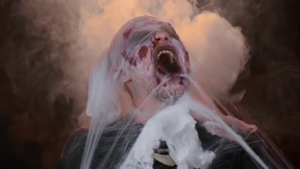 Frightening Man Halloween Zombie Bloody Wounded Hiding Behind Hands Blows Smoke From Nose and Mouth