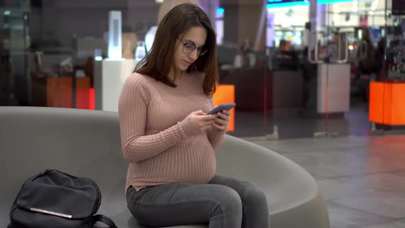 A Young Pregnant Woman in Glasses Sits with a Phone on a Bench