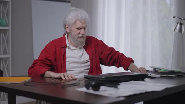 Senior Bearded Caucasian Disabled Man Closing Laptop Rolling Wheelchair to Window in Home Office