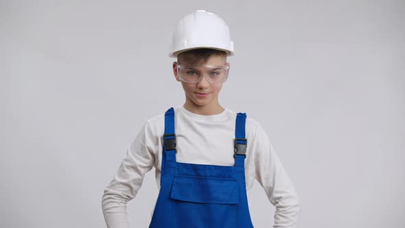 Smiling Charming Boy in Overalls Hard Hat and Eyeglasses Showing Peace Gesture Posing at White