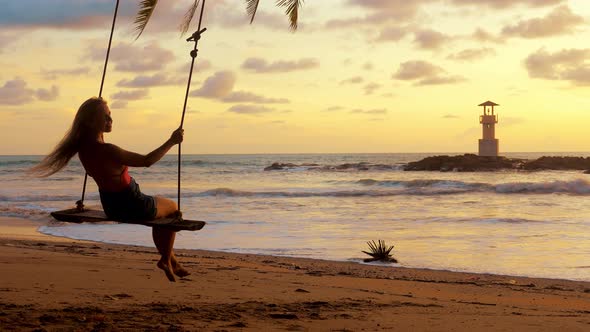 Young Smiling Woman Swinging on Rope Swing Suspended From Palm Tree at Sea Beach in Sunset Lights