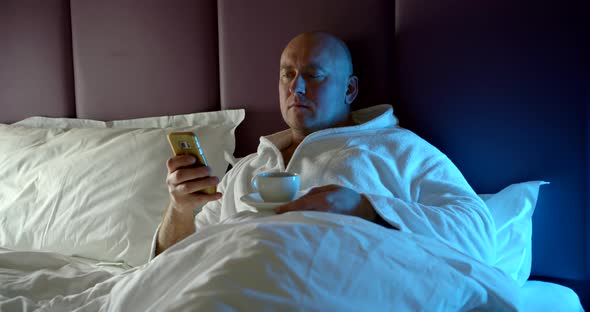 In the Evening in the Bed a Bald Adult Man Lies Under a White Blanket with a Cup of Tea and Watches