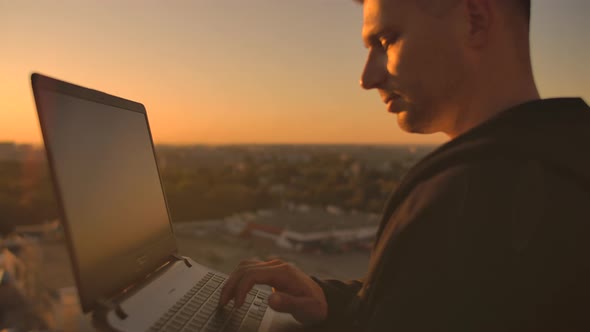 A Male Stockbroker Freelancer Stands on a Rooftop at Sunset with a Laptop and Types on a Keyboard