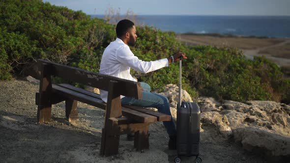 Wide Shot Side View of African American Man Sitting on Bench with Trolley Bag Looking at Turquoise