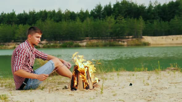 Young Attractive Man Sitting By the Campfire, Roast Marshmallows on a Stick. Against the Background