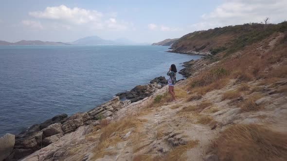 Aerial: a low altitude dolly in shot of a girl standing at a high hill looking at a rocky shore and