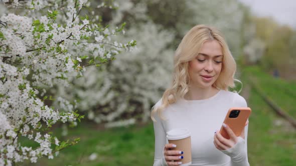 Happy Woman with Coffee and Mobile Walking in Spring Garden