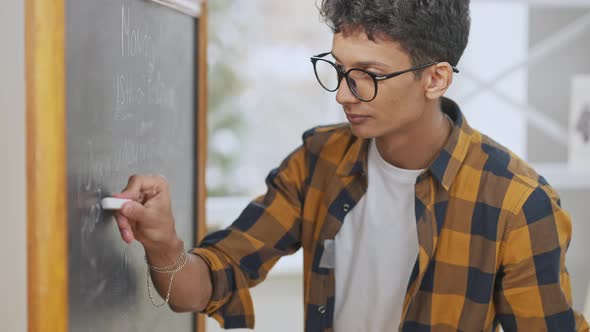 Portrait of Nerd Middle Eastern Teenager Writing with Chalk on Blackboard Looking at Camera and