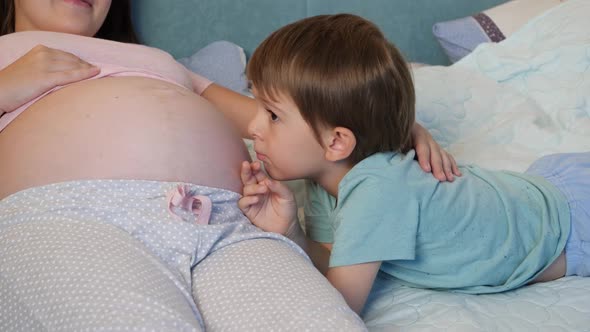 Portrait of Little Toddler Boy in Pajamas Lying with Pregnant Mother in Bed and Talking To Unborn