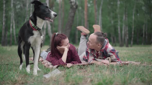 Happy Relaxed Caucasian Couple Lying on Blanket in Forest and Talking As Blurred Dog Standing at