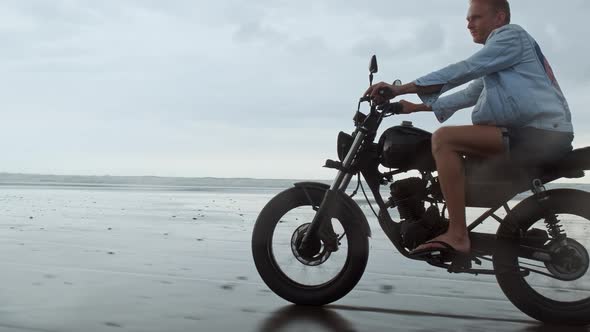 Young Handsome Hipster Man Riding Modern Custom Motorcycle Racer on the Black Sand Beach