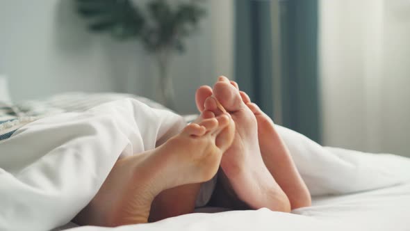 Human Bare Feet Touching Each Other in Bed Under White Blanket in Bedroom