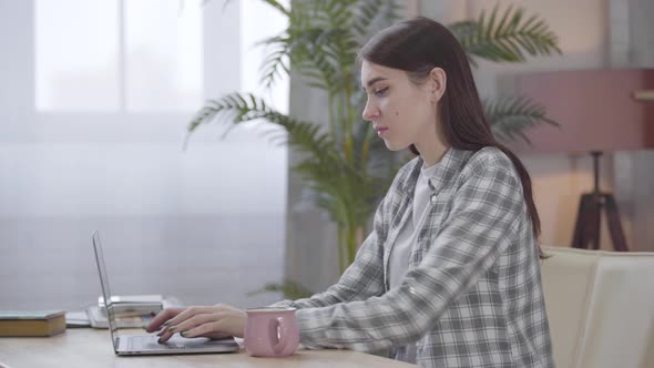 Side View of Young Caucasian Brunette Woman Putting Cut with Tea or Coffee at the Table and Typing