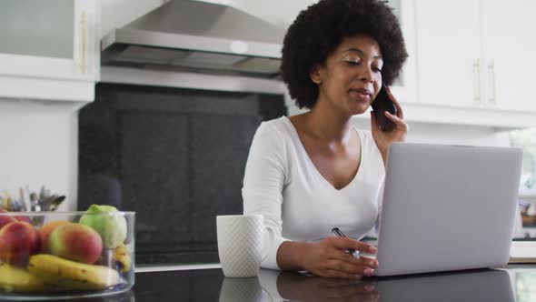 African american woman talking on smartphone and using laptop in the kitchen while working from home