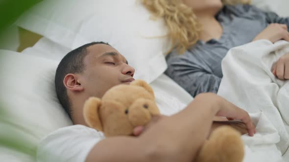 Closeup of African American Young Man Hugging Teddy Bear Sleeping in Comfortable Bed with Blurred