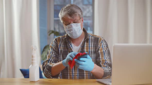 Senior Man in Mask and Gloves Disinfecting Laptop and Smartphone at Home