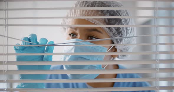 African Doctor in Protective Mask Gloves and Cap Looking Out Window Through Ajar Blinds