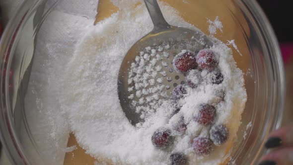 Slow motion overhead metal strainer sifting bright red cranberries and sugar in clear glass bowl on