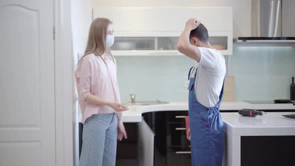 Side View of Young Caucasian Woman in Covid Face Mask Explaining Breakage To Carpenter in Apartment