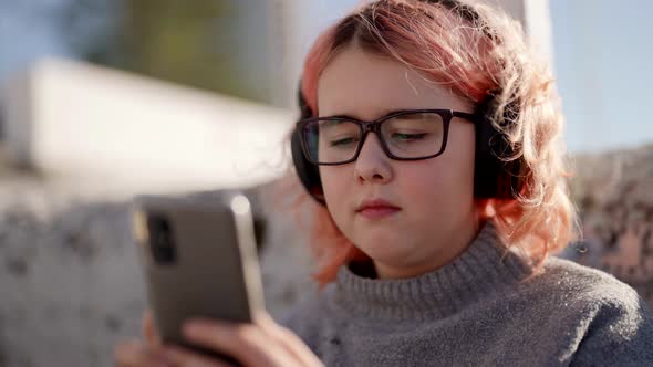 A Cute Teenage Girl in Windy Sunny Weather Sits Outside and Listens to Music with Headphones and